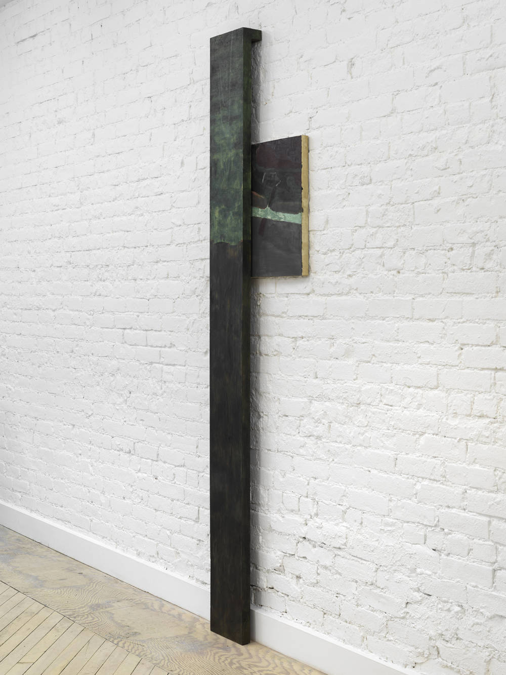 A vertical sculpture and painting rendered in a dark black and green palette installed against a brick gallery wall. 
