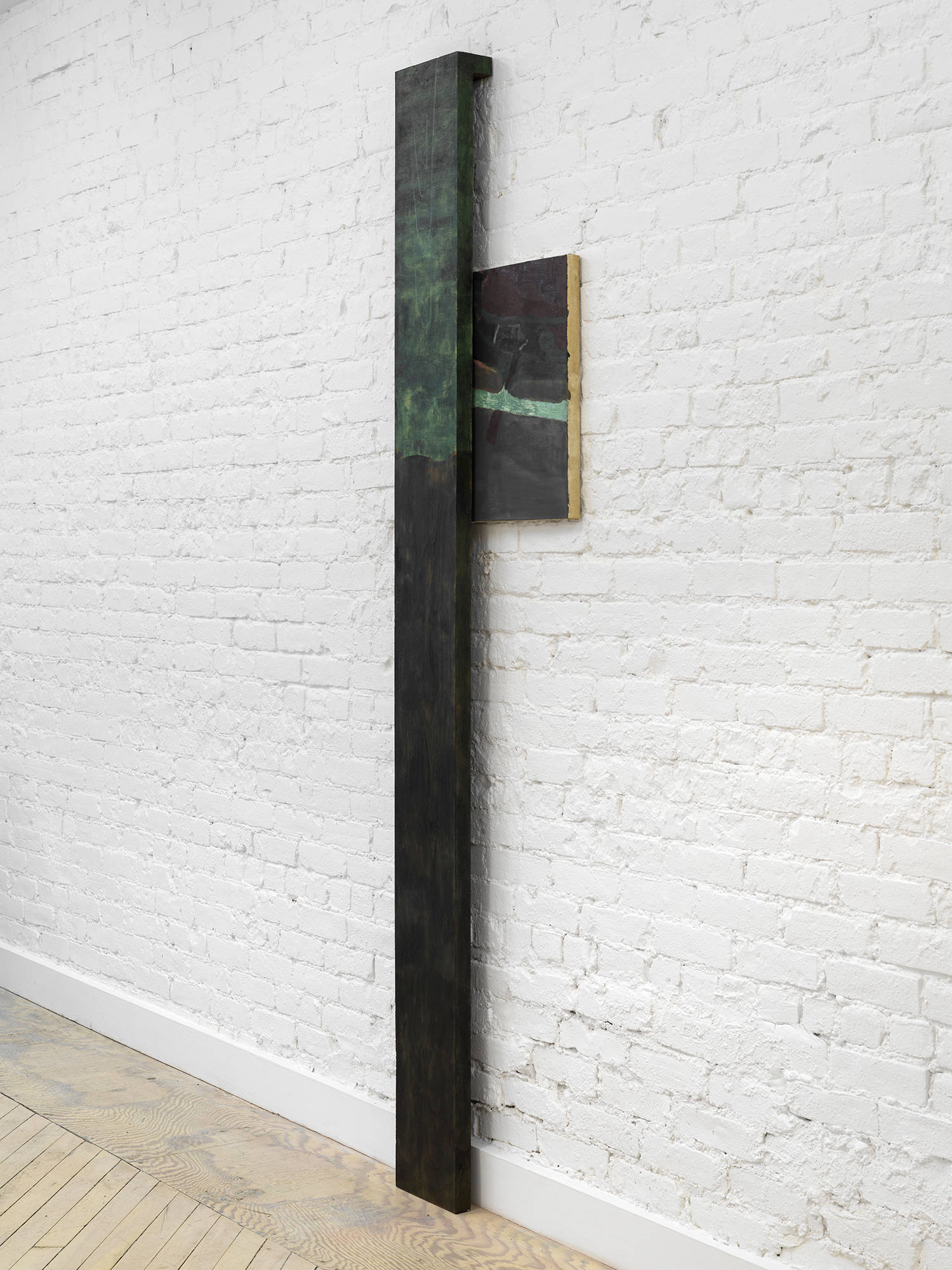 A vertical sculpture and painting rendered in a dark black and green palette installed against a brick gallery wall.