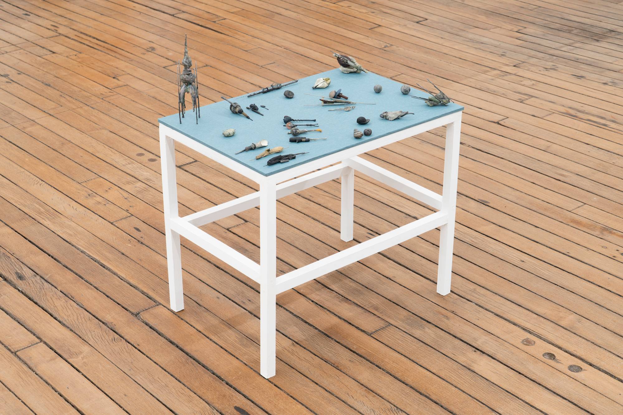 A small, wood table, painted white, with blue fabric top holds an array of abstract miniature sculptures