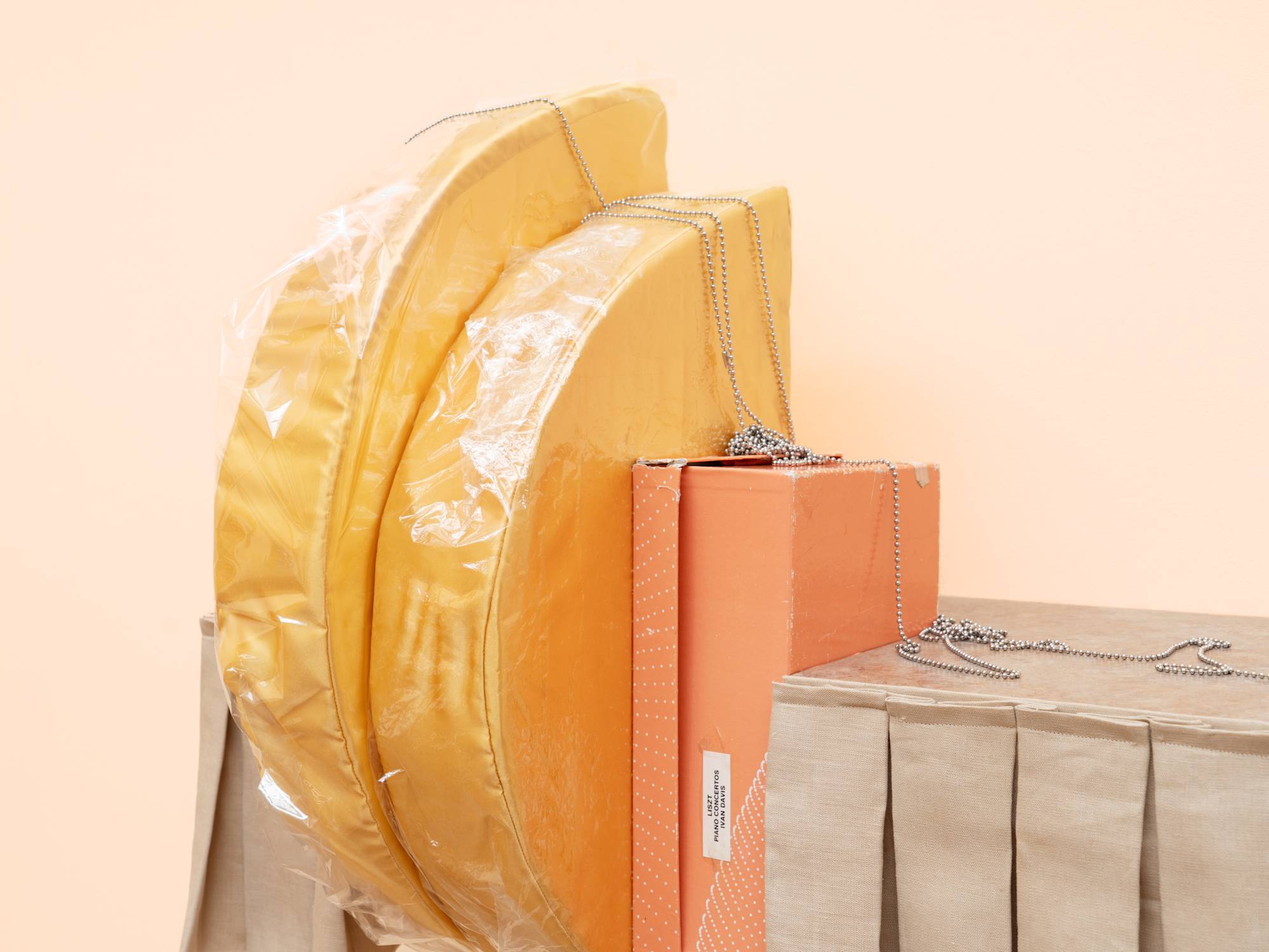 Detail of a sculptural shelf hanging on a peach colored wall. The shelf is lined with a short, beige, pleated curtain that is parted to the left of center by two vertical, yellow cushions, wrapped in cellophane, and an orange shoe box. A small picture hangs from the right side of the shelf by a ball chain.