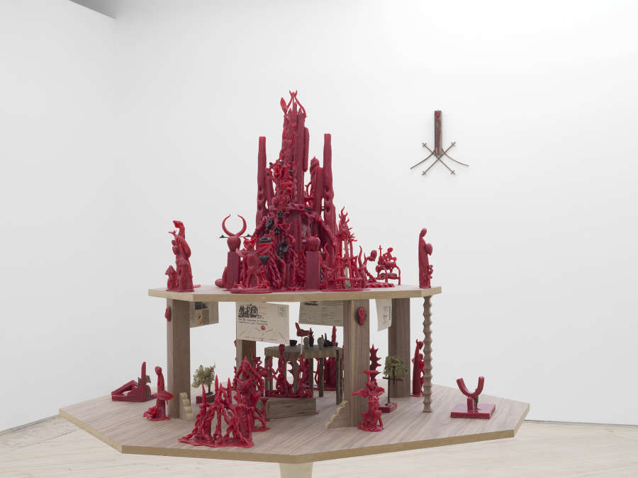 Corner installation view of Harry Gould Harvey IV sculpture made of smaller red wax sculptures resting on a tiered wooden platform on top of a tulip table base, with one small bent wood sculpture hanging on the wall behind it.