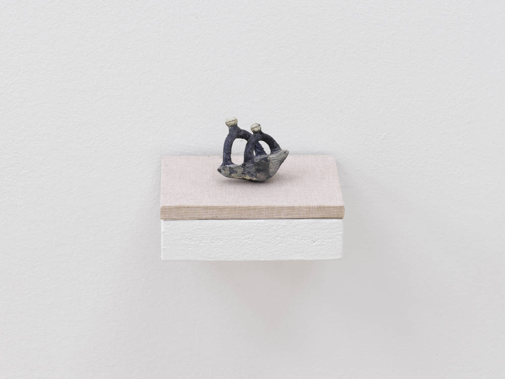 A small abstract sculpture resembling a kind of prehistoric instrument or piece of jewelry. The object is primarily blue and gray and resting on a shelf with a tan base attached to a white wall.