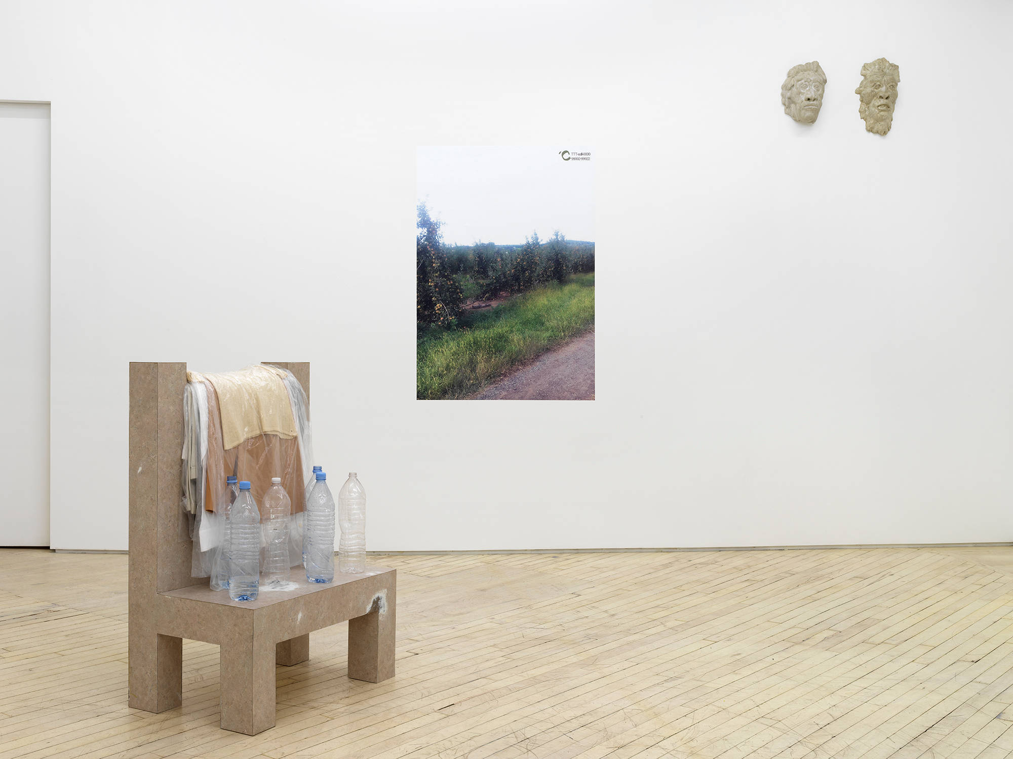 Gallery shot of three artworks. One is a sculptural piece, resembling the form of a chair with empty waterbottles on top. Another, an image of a nature scene. And last, two masks hanging up on the wall.