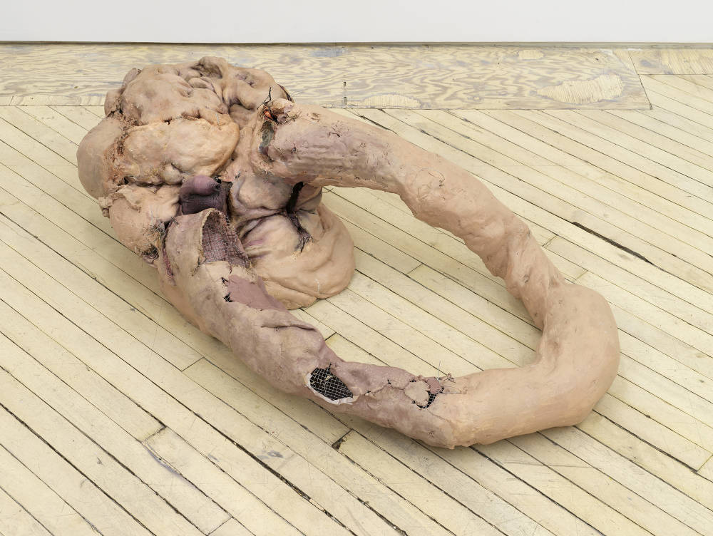 In a gallery space, an abstract sculpture rests on the floor resembling human flesh.