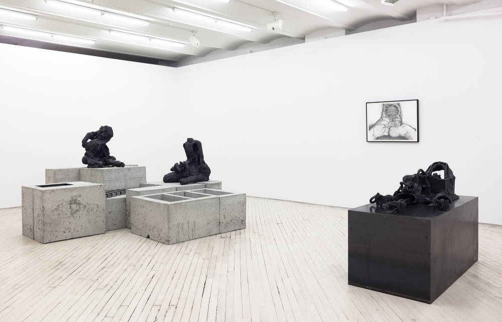 Installation view of 2 sculptures and at the back right a black framed black and white drawing. The sculpture at left is larger and appears to be made of a grey concrete base comprising many differently shaped and sized blocks, atop which sits two black, hunching figures. At right is a sculpture that is mostly black, featuring a sleek large rectangular base and a more biomorphic black object on top.