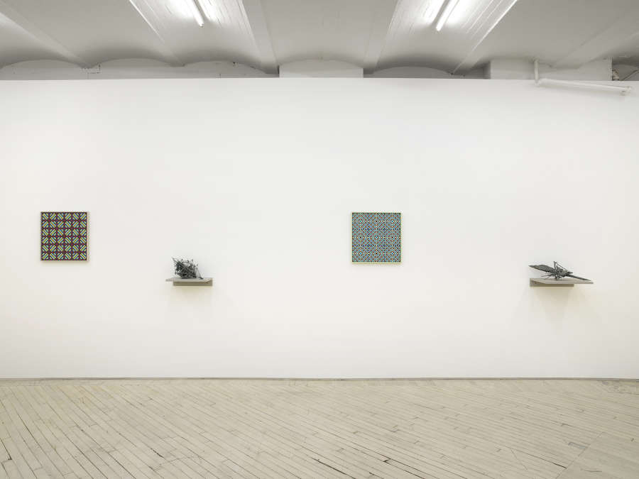 On a long white gallery wall, two square paintings depicting a grid of crisscrossing lines rendered primarily in blues, purples, and greens. separating the paintings are two abstract sculptures resembling archaic amalgamations of organic material. The sculptures are resting on top gray shelves attached to the wall lower than the paintings. 