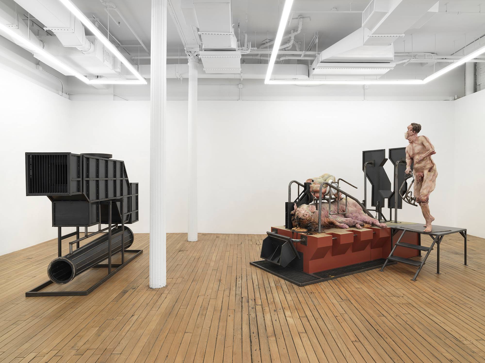 In a large gallery space, two monumental sculptures are spaced apart. The sculpture on the left is a large steel structure. To the left a combination of abstract body parts and a form resembling a pig.