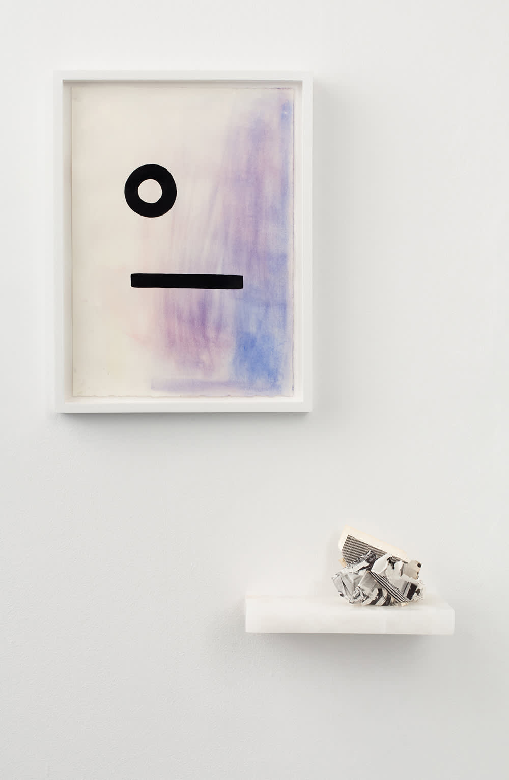On a gallery wall, a drawing in a white frame hangs above a shelf housing a small sculptural object.