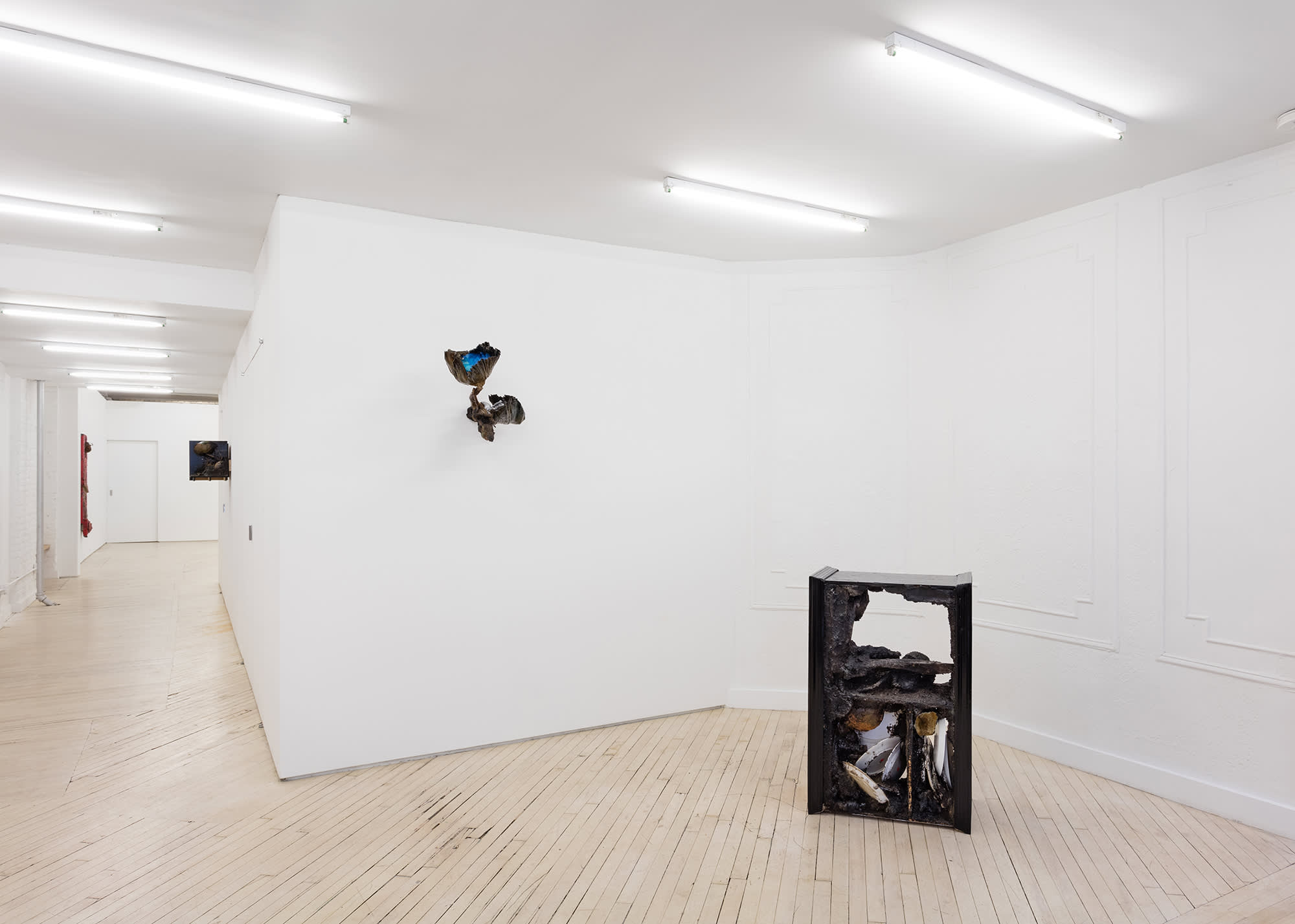 Installation view of an exhibition by Brandon Ndife. In a gallery there are two sculptures, one hanging on the wall and one in front of it on the floor. Both are brown/black and covered in dirt and debris. The wall sculpture appears like a plant and a sconce. The floor sculpture looks like a piece of furniture. You can see down the hallway that there are additional sculptures hanging on the walls.