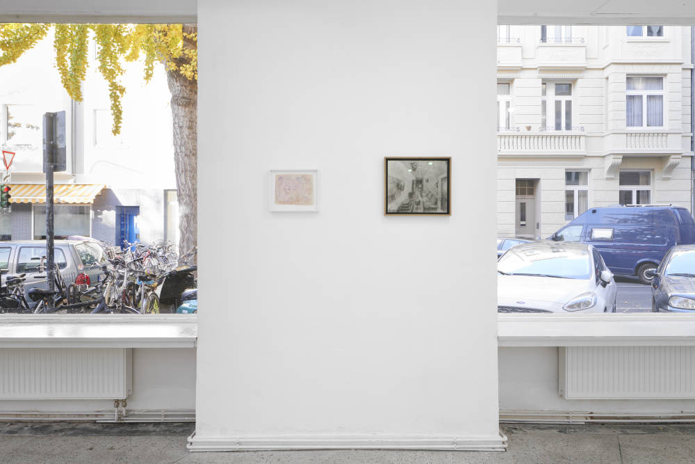 In a gallery space, two small works on paper framed and installed between two large windows.