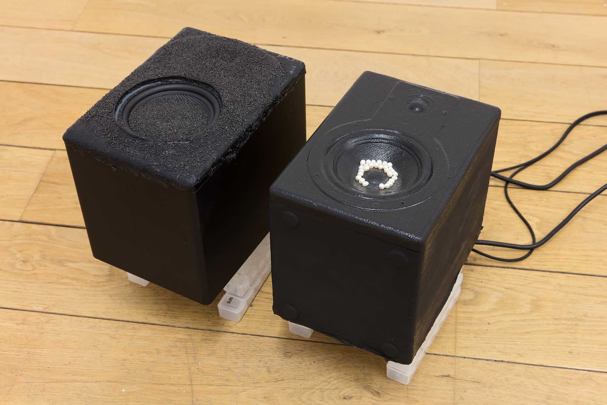 Birds eye view of two black speakers on a blond wooden floor with black cables trailing off out of the frame on the right. In one of the speakers is black sand in the cone of the speaker and one has white pearls inside.