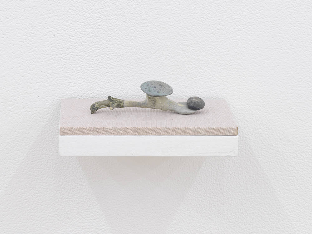 A small abstract sculpture resembling a kind of fossil, bone, or combination of found organic materials. The object is primarily gray and resting on a shelf with a tan base attached to a white wall.
