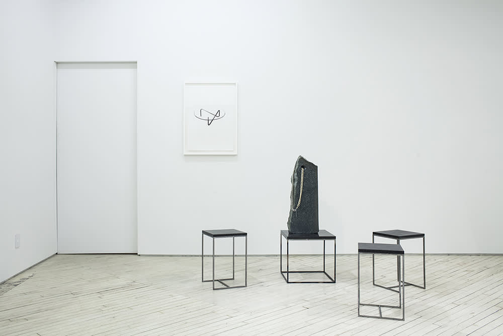 Installation view of a black ink drawing on white paper framed in white at the back wall, in front of which is a set of steel and black marble square stools, one of which is supporting a large marble stone perforated by a hold, through which a long golden chain is threaded.