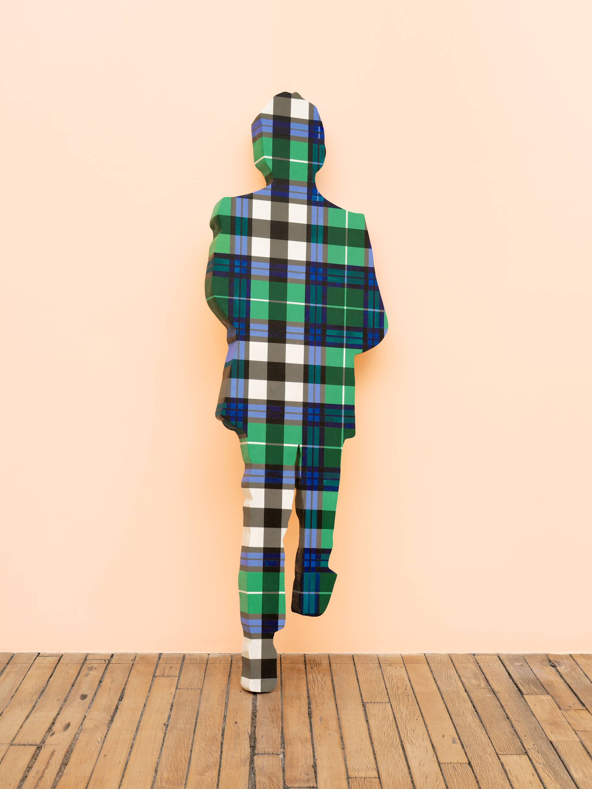 A life-sized human figure painted in a blue and green plaid pattern leans against a peach wall in an art gallery.