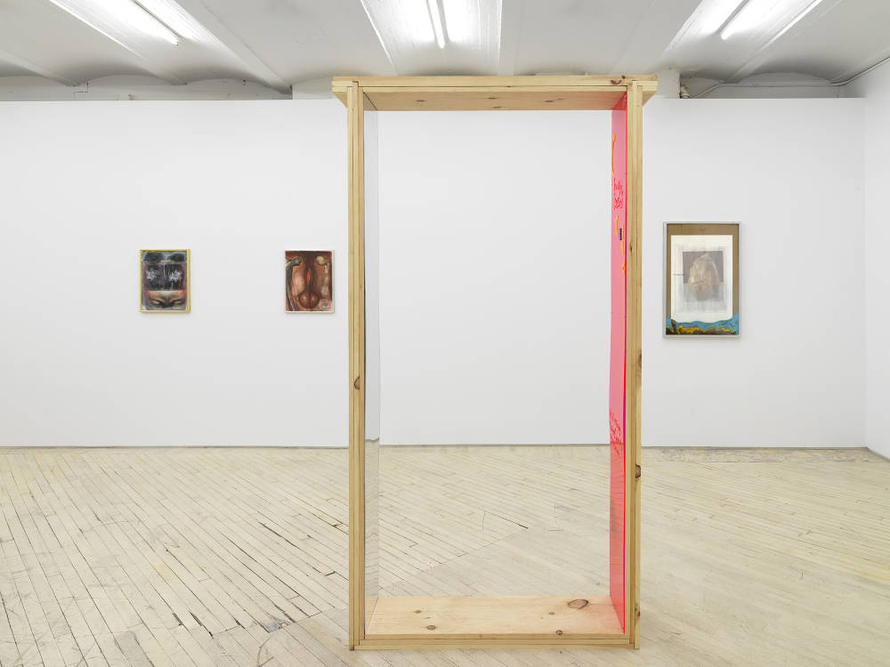 In a gallery space, two small paintings on the left. In the center a large wooden rectangular sculpture with one mirrored side and another in pink plastic. To the right a large photograph with a brown border. 