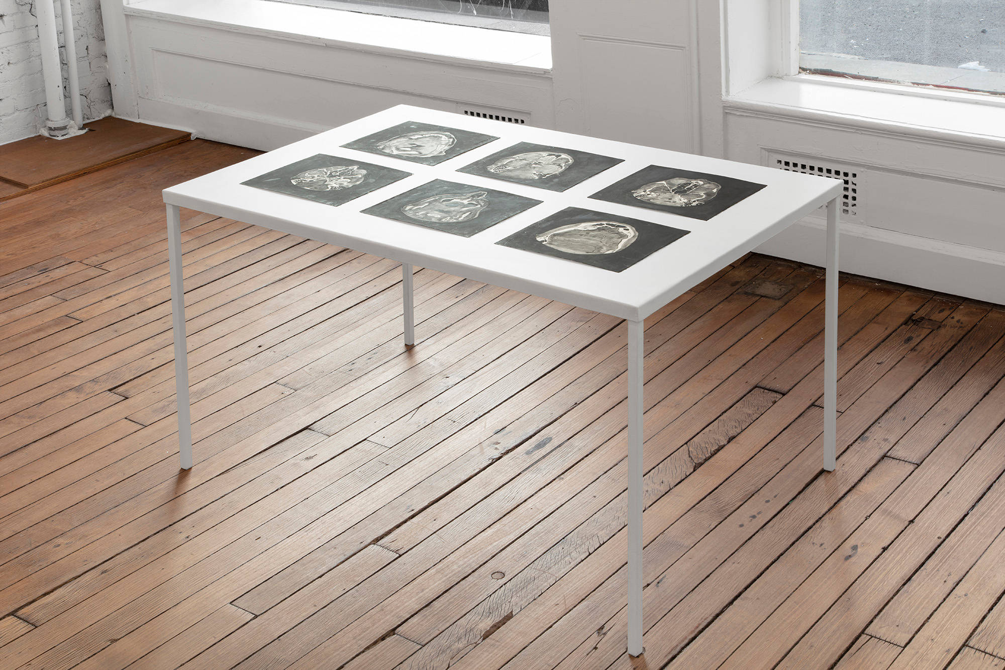 A steel table, painted white, with four legs stands on a wooden floor near two large windows. The table holds six liquid graphite drawings depicting MRI scans of a sinus cavity.