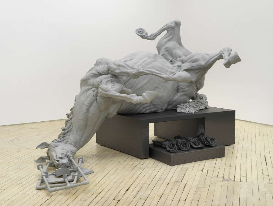 In a gallery space, a large sculpture of a fallen horse resting on top of an iron base. 