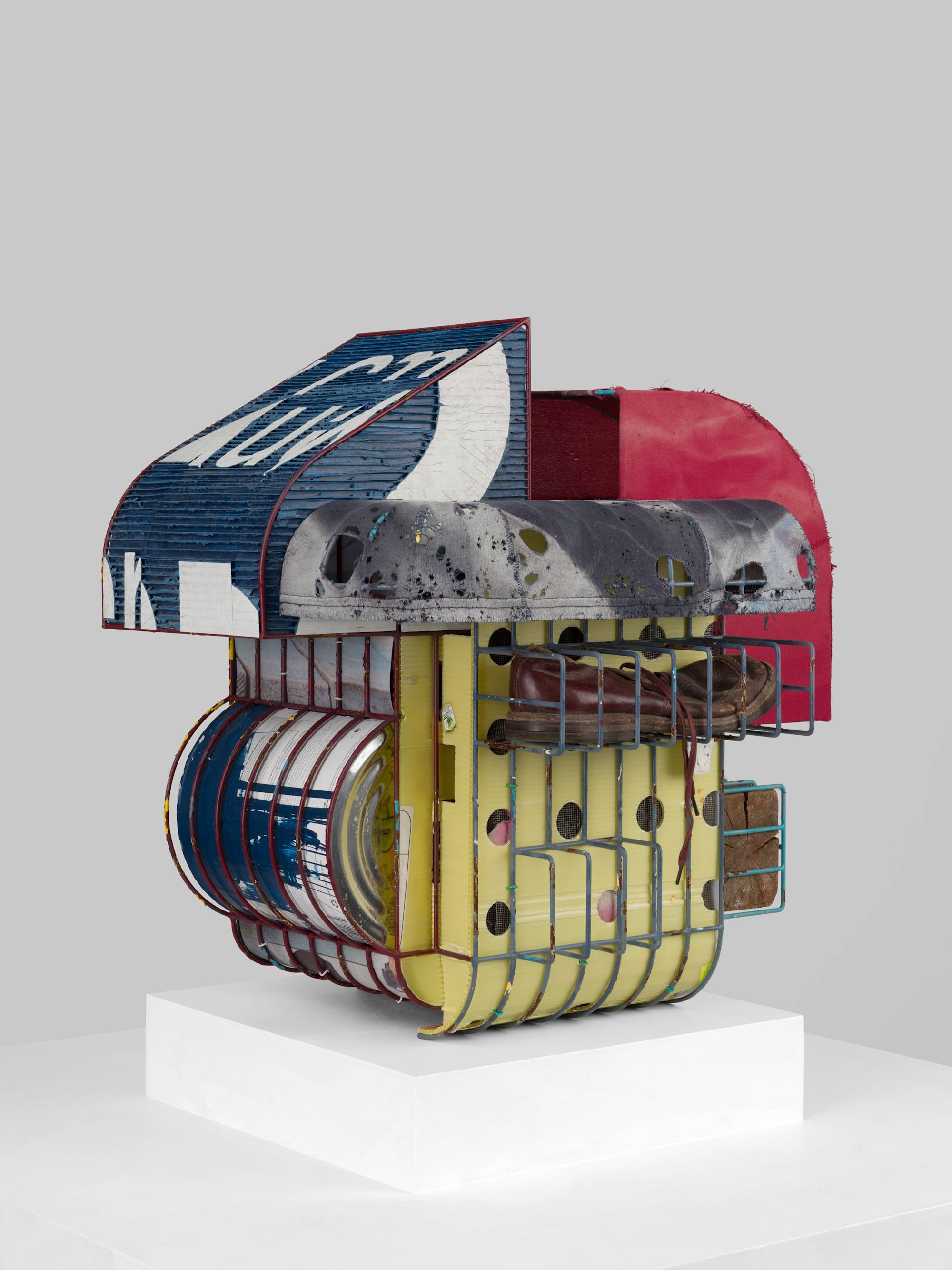 A cage-like colorful steal wire sculpture, wrapped with several tattered fabrics to resemble various awnings, and embellished with an old shoe, a twig, printed coroplast signage, an old blue paint can, a lock, a paper tag, and a wooden block sits on a white pedestal against a white wall.
