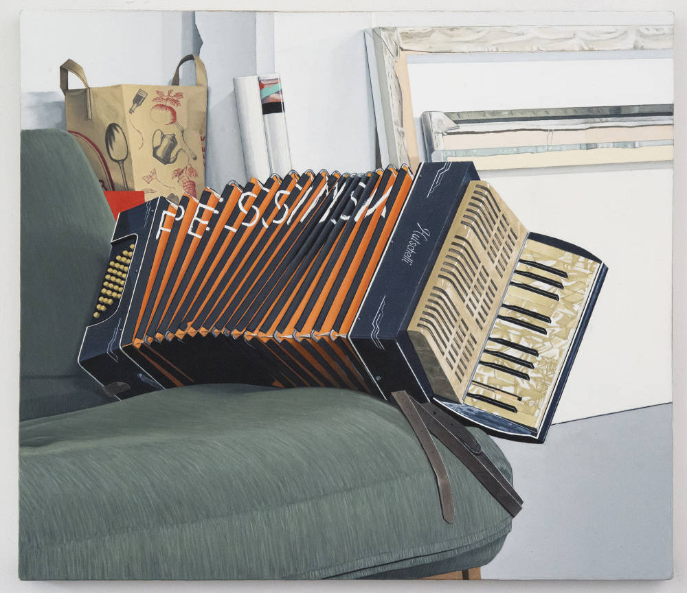 Oil on canvas painting depicting an accordion sitting on a green futon couch, inside the baffles of the accordion is written the word "Pessimism." In the background there is a brown paper bag from Trader Joe's and several paintings leaning face towards a wall.