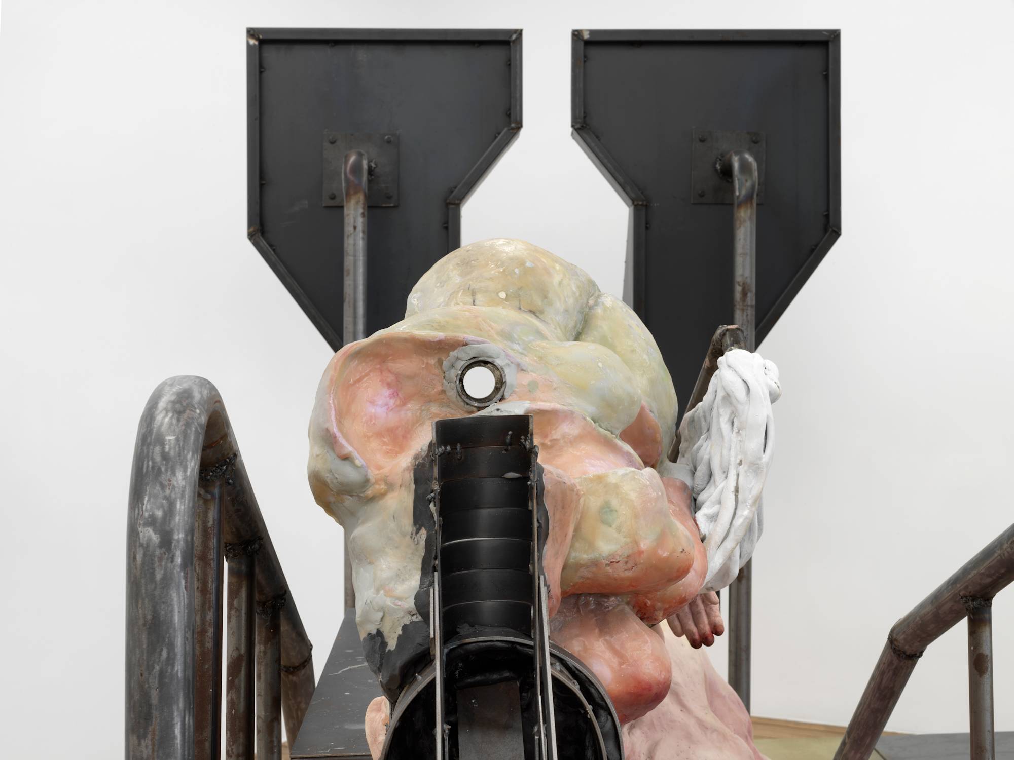 Close up image of a piece of metal resembling a stairwell handrail and a larger steel structure in back. An abstract pink sculpture is fixed in the middle.