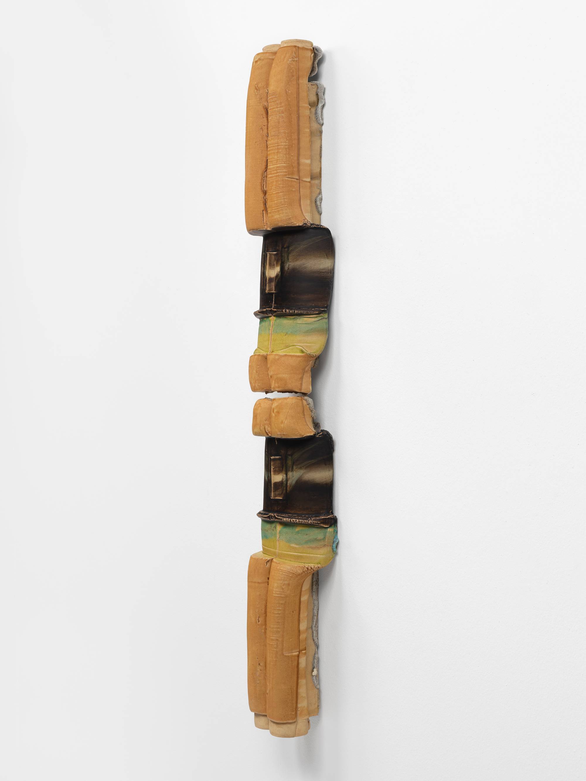 Three quarter view of a large, vertically oriented, 2-piece sculpture, featuring cylindrical elements, in hues of mustard, brown, and green, hanging on a white wall.