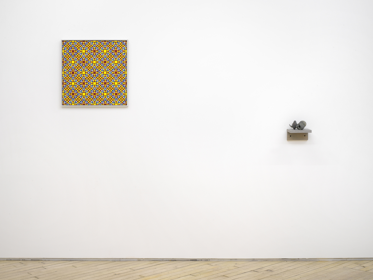 On a white gallery wall to the left a painting consisting of a repeating geometric pattern of criss-crossing lines. The dominant colors are yellow, red and orange in a gray frame. To the right is small abstract sculpture resembling some combination of archaic materials such as bones. The object is resting on a gray shelf hung lower than the painting. 
