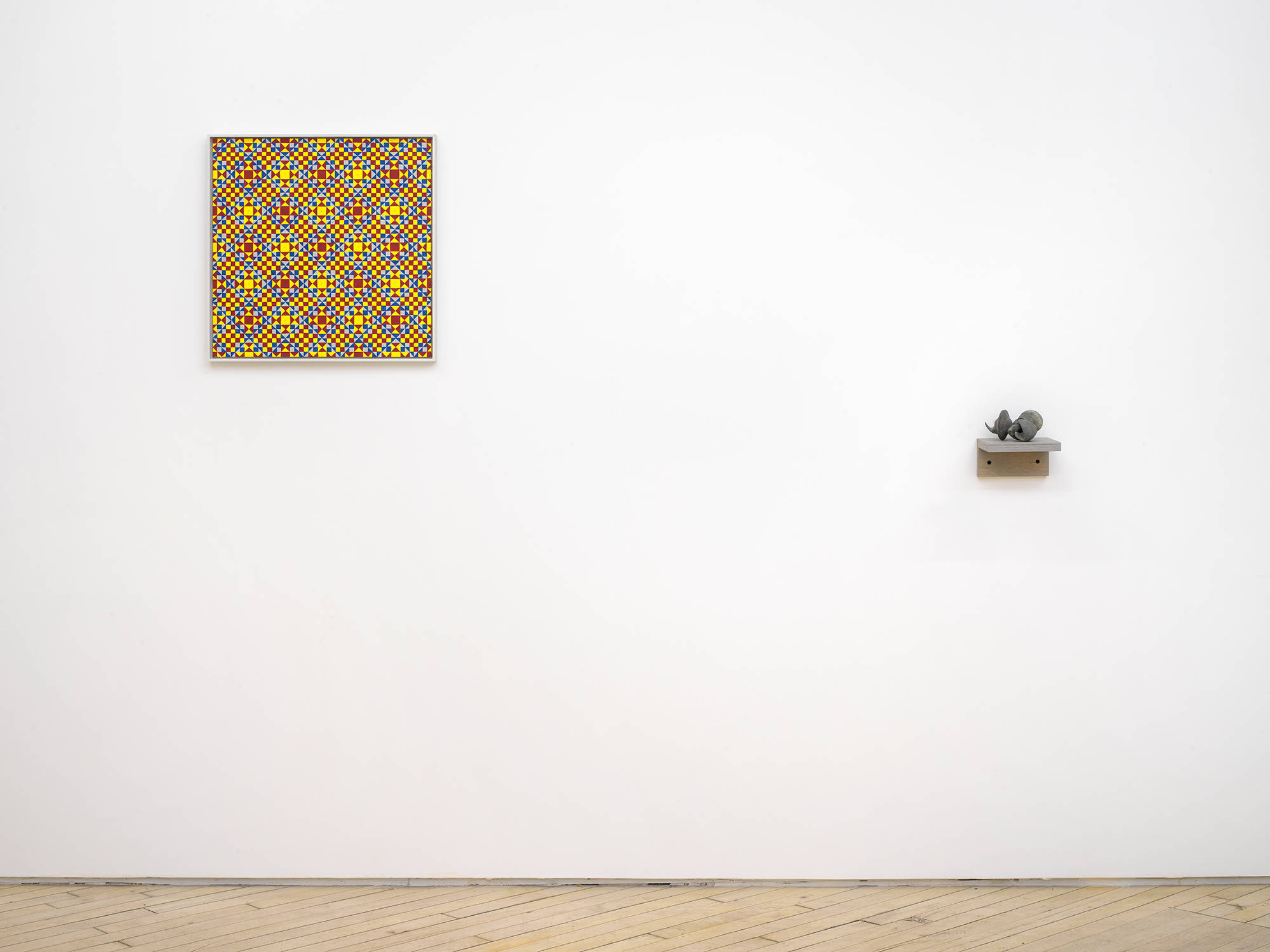 On a white gallery wall to the left a painting consisting of a repeating geometric pattern of criss-crossing lines. The dominant colors are yellow, red and orange in a gray frame. To the right is small abstract sculpture resembling some combination of archaic materials such as bones. The object is resting on a gray shelf hung lower than the painting.