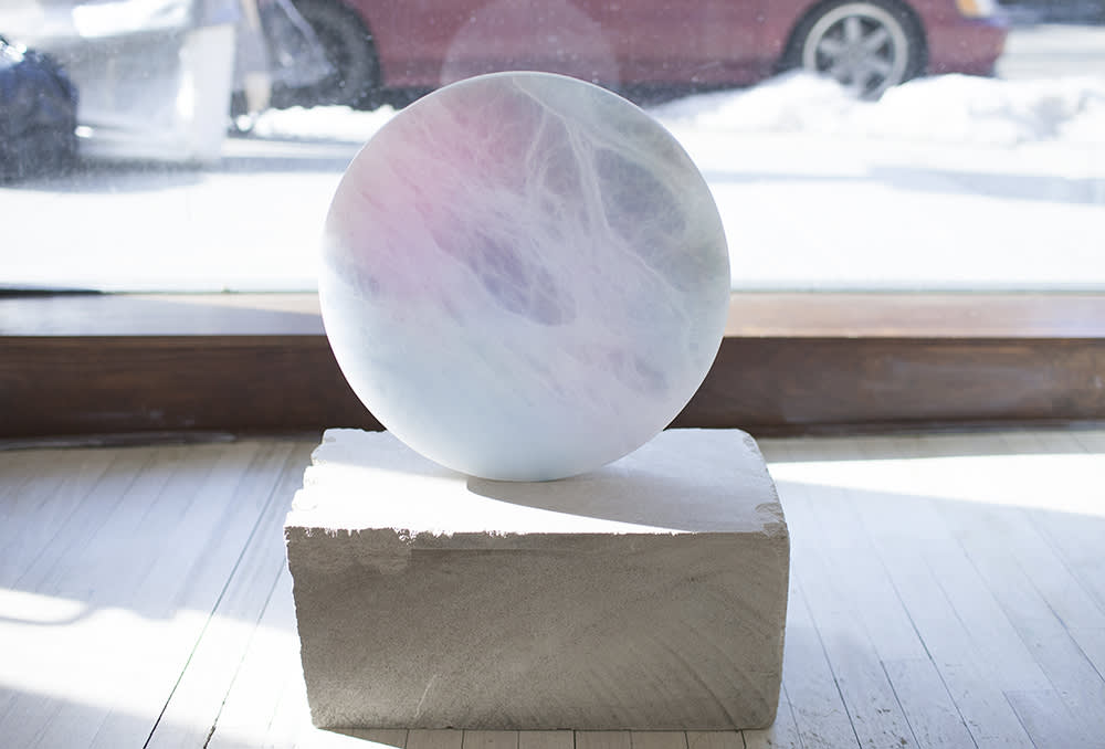An alabaster disc rests on top of a stone plinth, placed in front of a large floor window looking out onto a snowy street with a red car parked at the distance. The disc is lit from behind with the daylight and has an airbrushed spray of pink and blue watercolor on the surface.