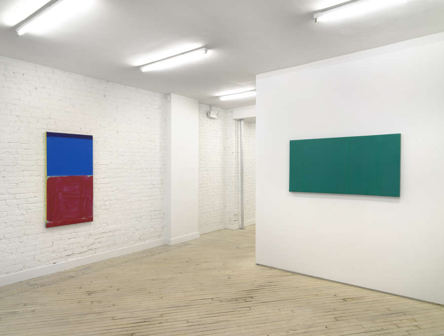 In a gallery space, a vertical painting consisting of a top blue portion and a bottom red portion hung on the left wall. On the right wall is a horizontal monochromatic green painting. 