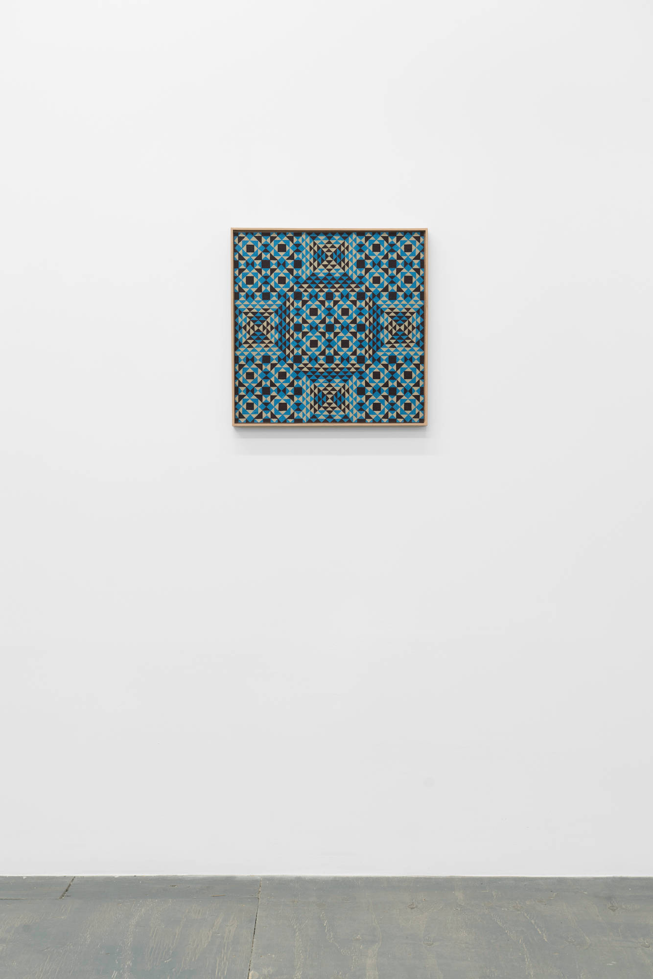 A colorful, symmetrical, geometric painting on board consisting of a pattern of repeating diamonds and triangles of tan, brown, and blue in a tan artist frame sits on a white wall above a grey wood floor.