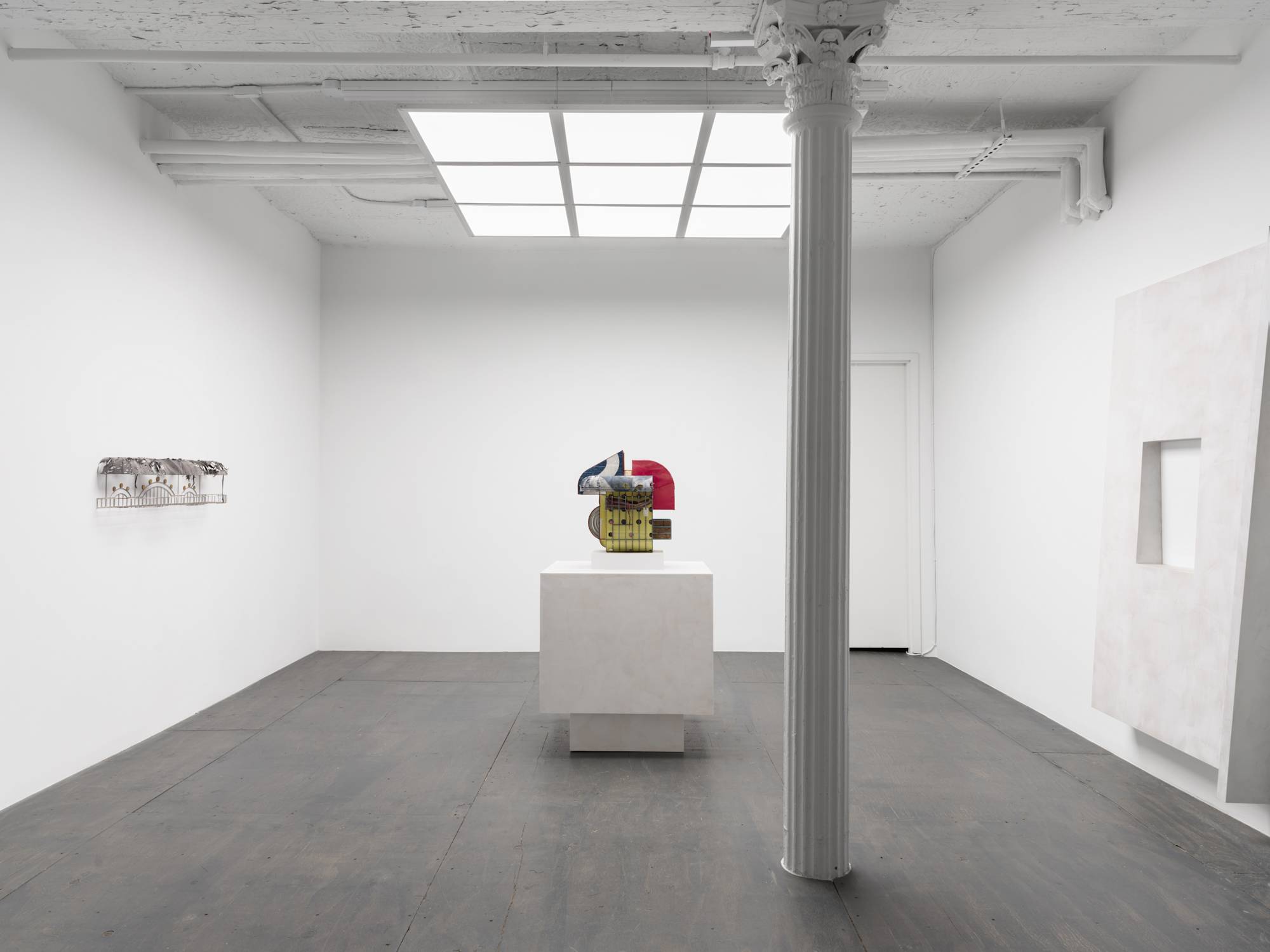 A white walled gallery with grey floor and white column lit by an LED light fixture that resembles a skylight. Three sculptures are featured in the gallery. Two on opposing walls and one standing on a pedestal in the center of the room.