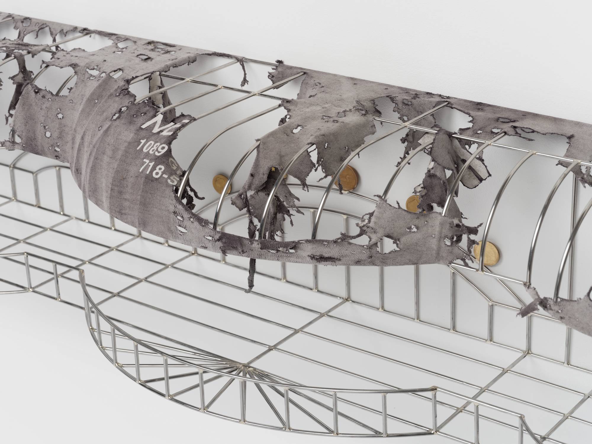 Detail of a cage-like chrome sculpture, wrapped with a tattered grey t-shirt to resemble an awning, and embellished with soldered on gold-colored coins.