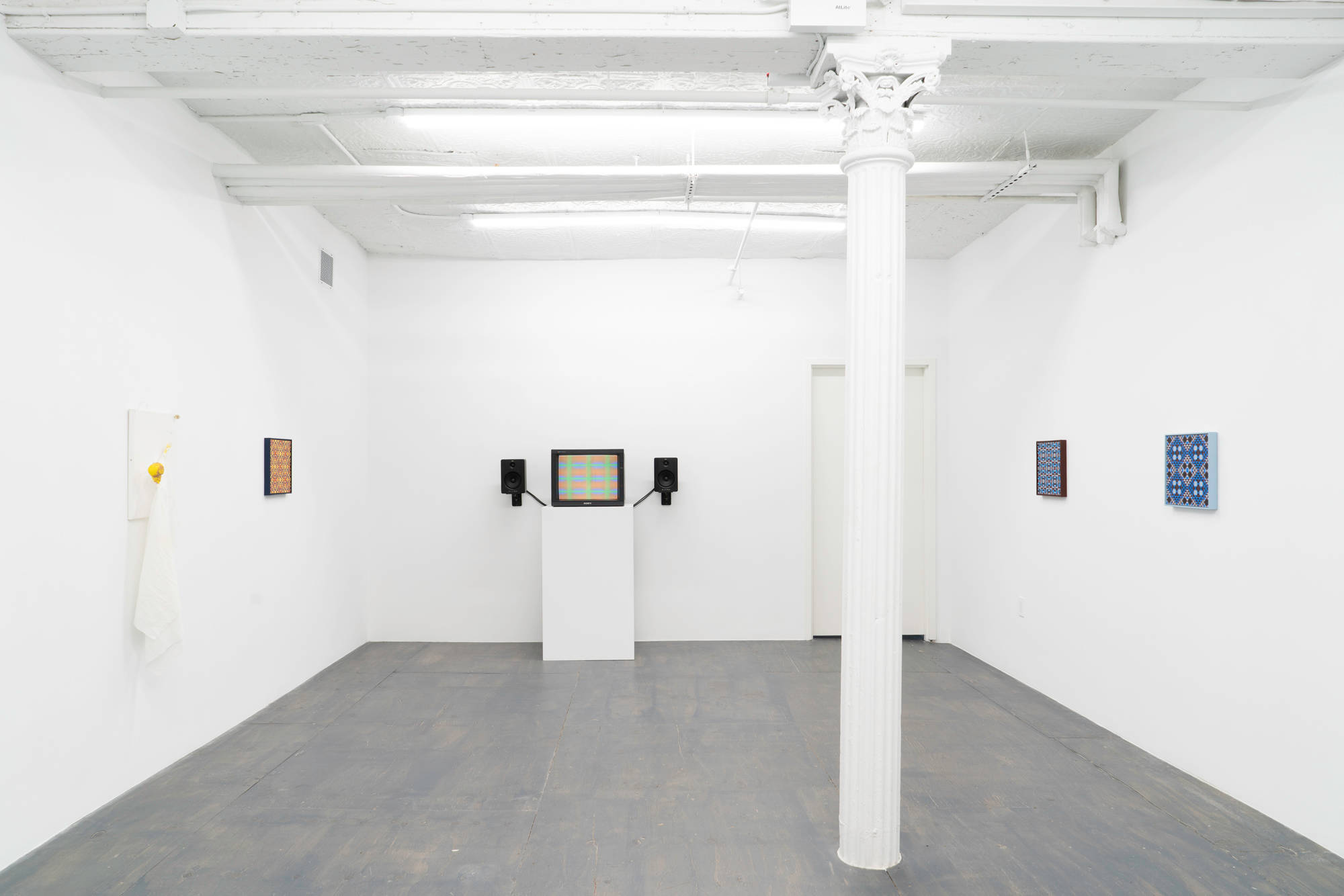 A white painting with a puppet-figure adhered to it hangs next to a geometric painting consisting of a pattern of repeating diamonds and triangles hung along a gallery wall. To the right a tv monitor displaying a still image blurred colors in a grid pattern taken from an animation, resting on top of a white pedestal with black speakers mounted to the wall on either side. To the right of the tv are two geometric paintings consisting of a pattern of repeating diamonds and triangles.