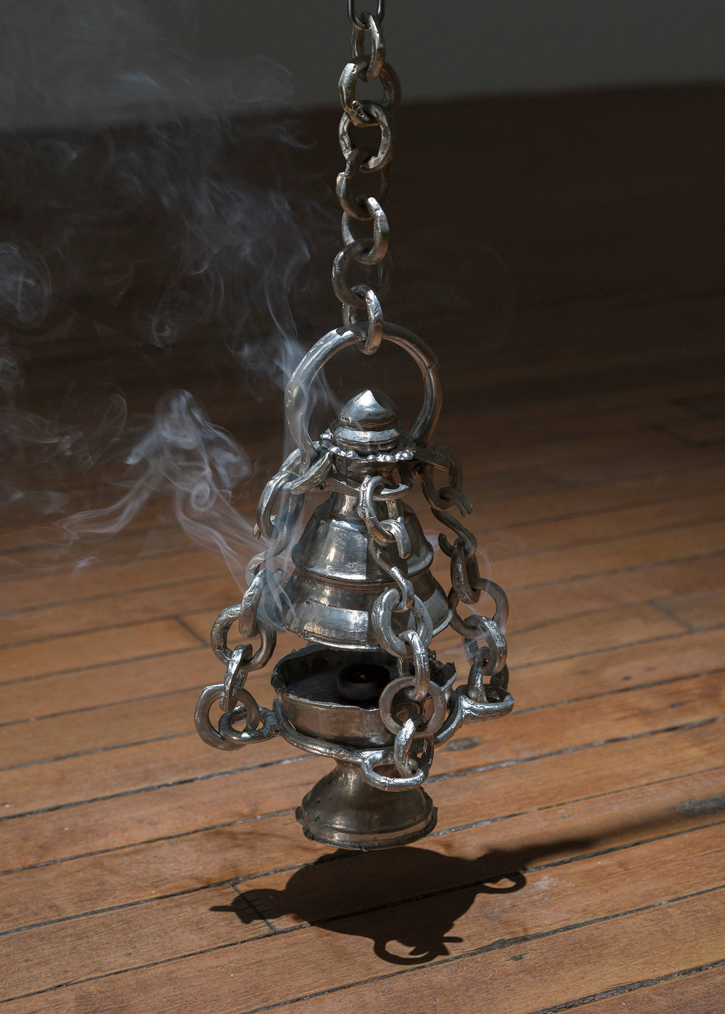 A close up view of a pewter thurible burning incense that hangs on a chain from the ceiling of a dark art gallery.