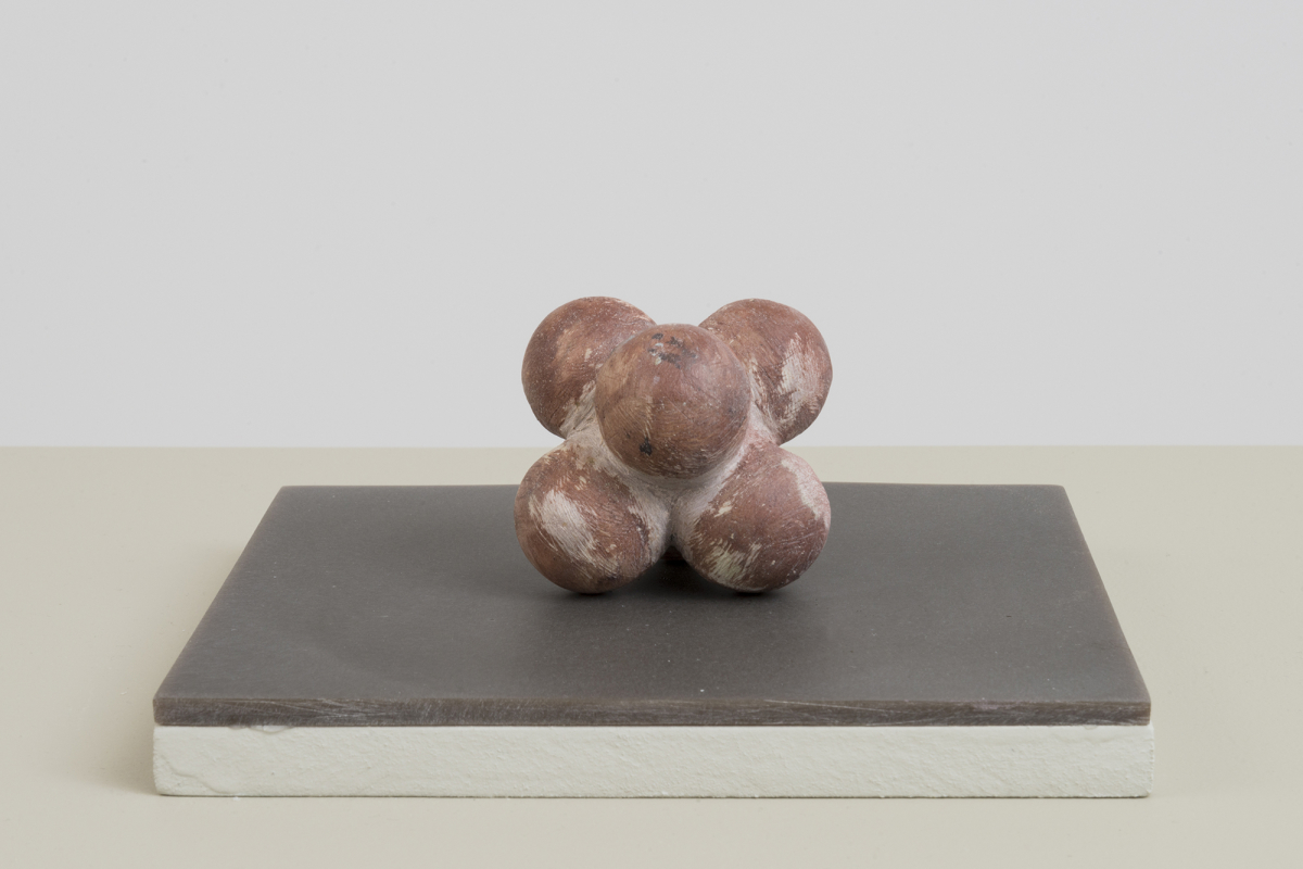 A handmade sculpture resembling a connected bunch of round bulbs resting on top of a brown-tinted plinth with a white wall partially in view. 