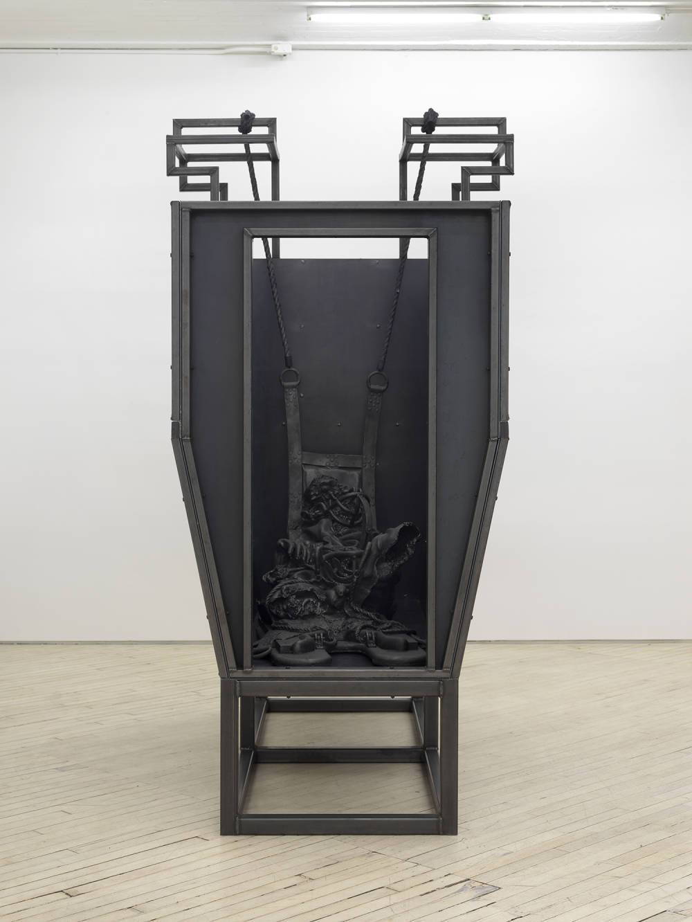Installation view of a metal sculpture resembling a cattle hold, containing sculptures made of cast iron depicting a decaying figure.