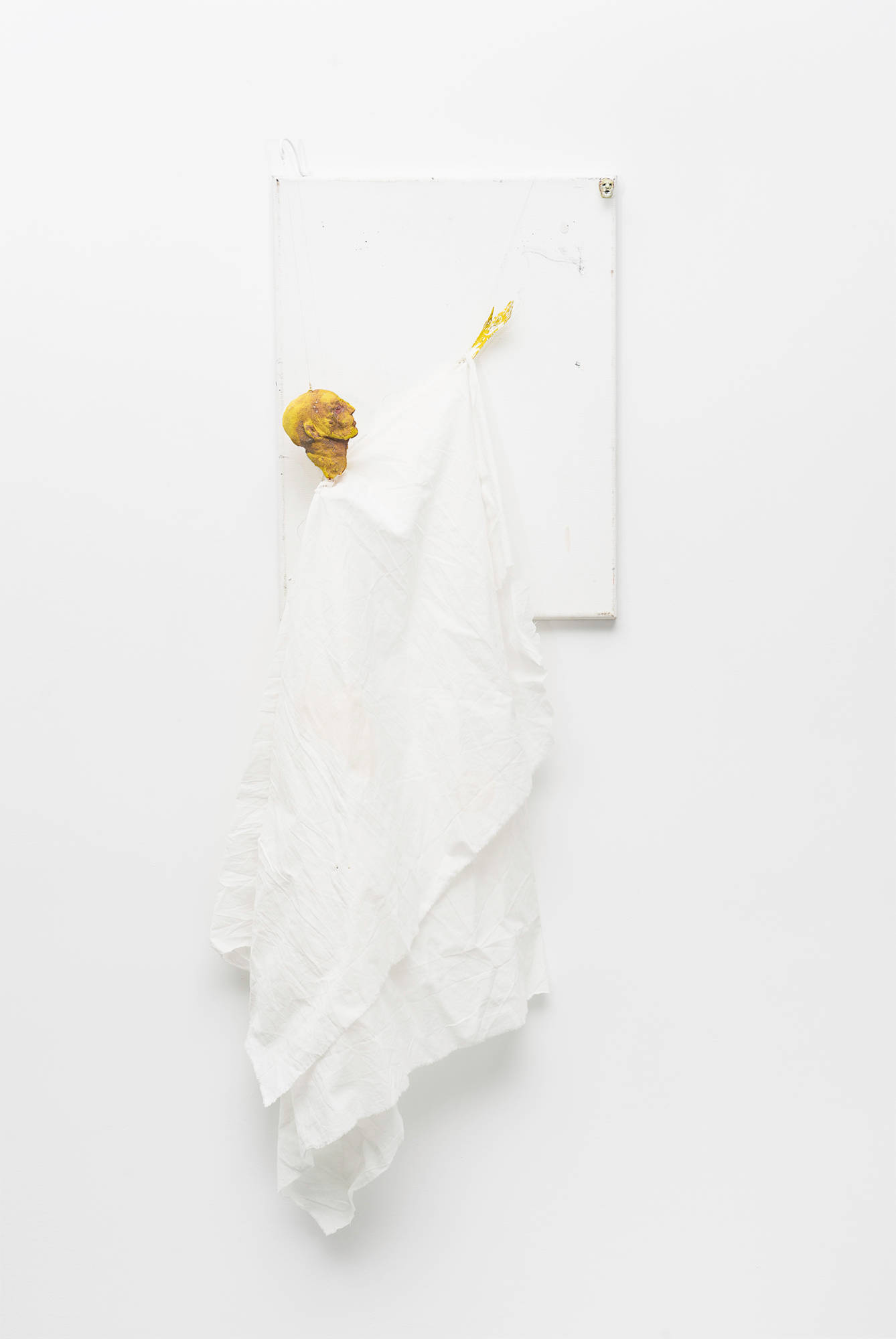 A white painting with a puppet-figure adhered to it hangs vertically along a white gallery wall.