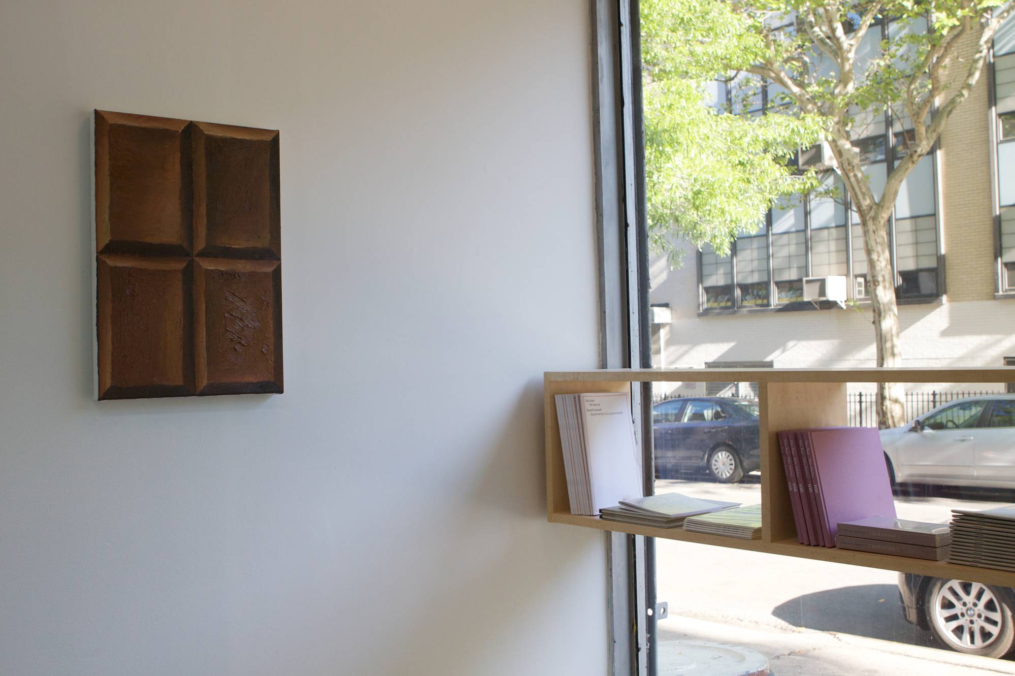 Image of a Viktor Kopp painting of 4 squares of brown chocolate in a window with a bookshelf.