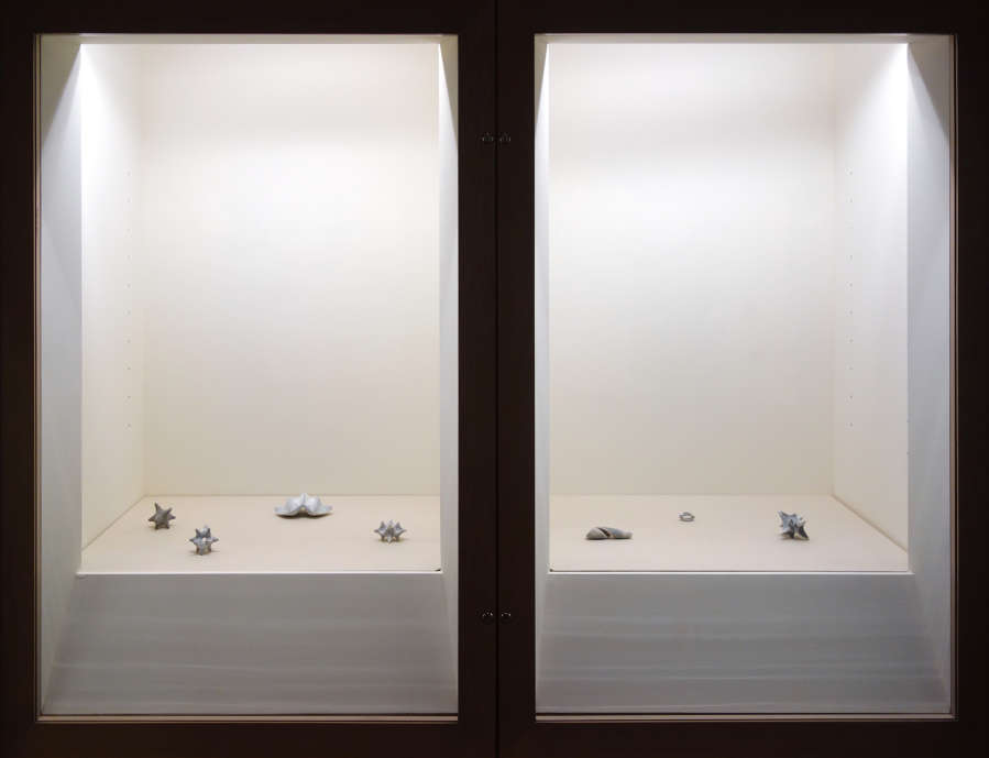 In two glass vitrines, several miniature abstract sculptures are arranged with varying amounts of space between the objects. 