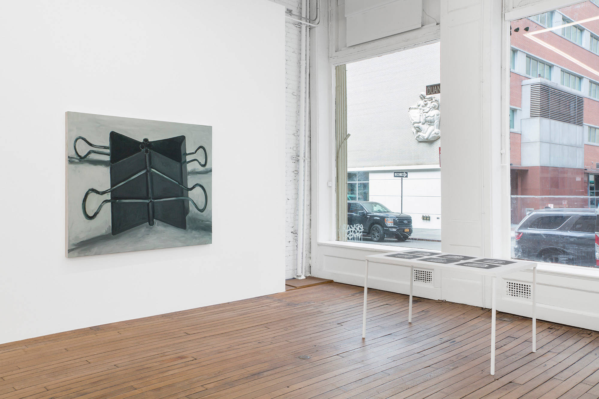 This image of the North West corner in the upstairs of the gallery contains a painting of binder clips on the end of the west wall and a table of six graphite drawings on paper laid on a table near the windows of the North wall.