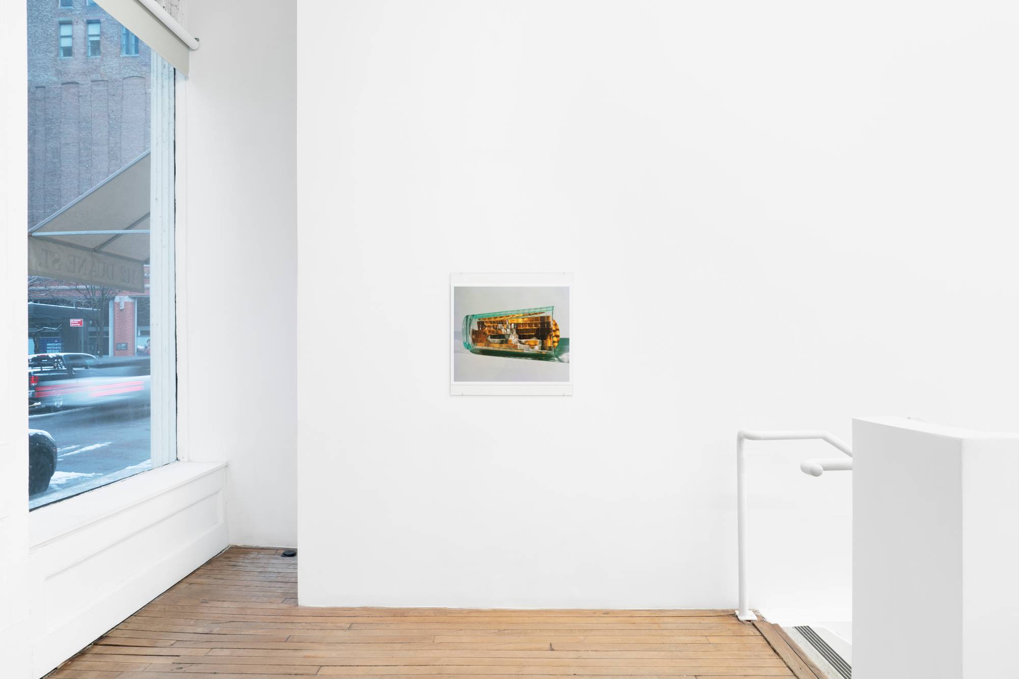 A photographic print of a blurred yellow object, resembling an ear of corn, slightly jutting out from inside a checkered glass cup hangs above a wood floor in an art gallery. The street can be seen from a window to the left and stairs descend to the right out of frame.