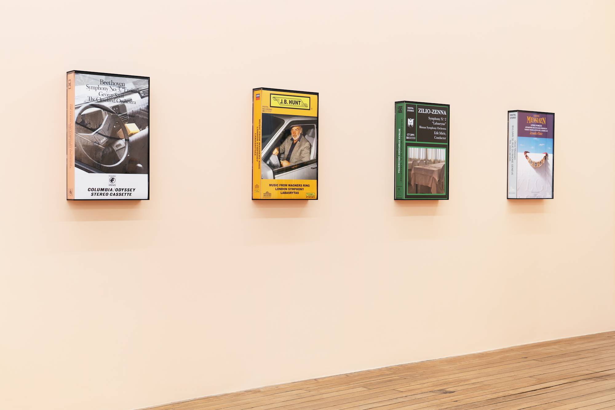 Four large, fake audio cassette cases featuring various artworks are displayed on a peach-colored wall in a gallery with wooden floors.