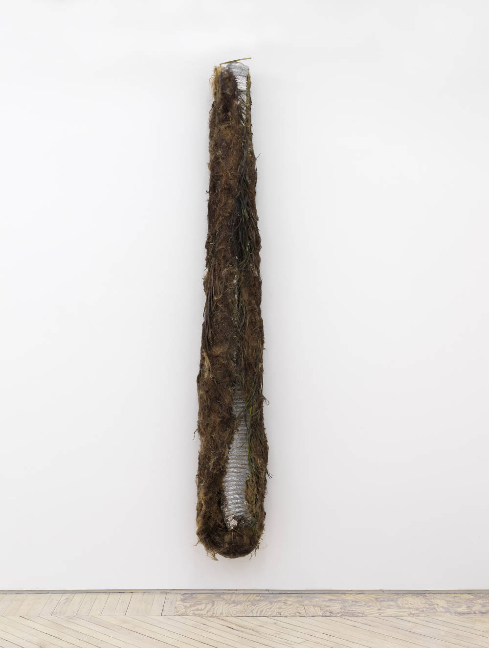 Image of a sculpture by Brandon Ndife showing a silvery tube of HVAC duct hung vertically on the wall. The ductwork is covered with a brown furry vegetal substance similar to a Ghillie Suit.