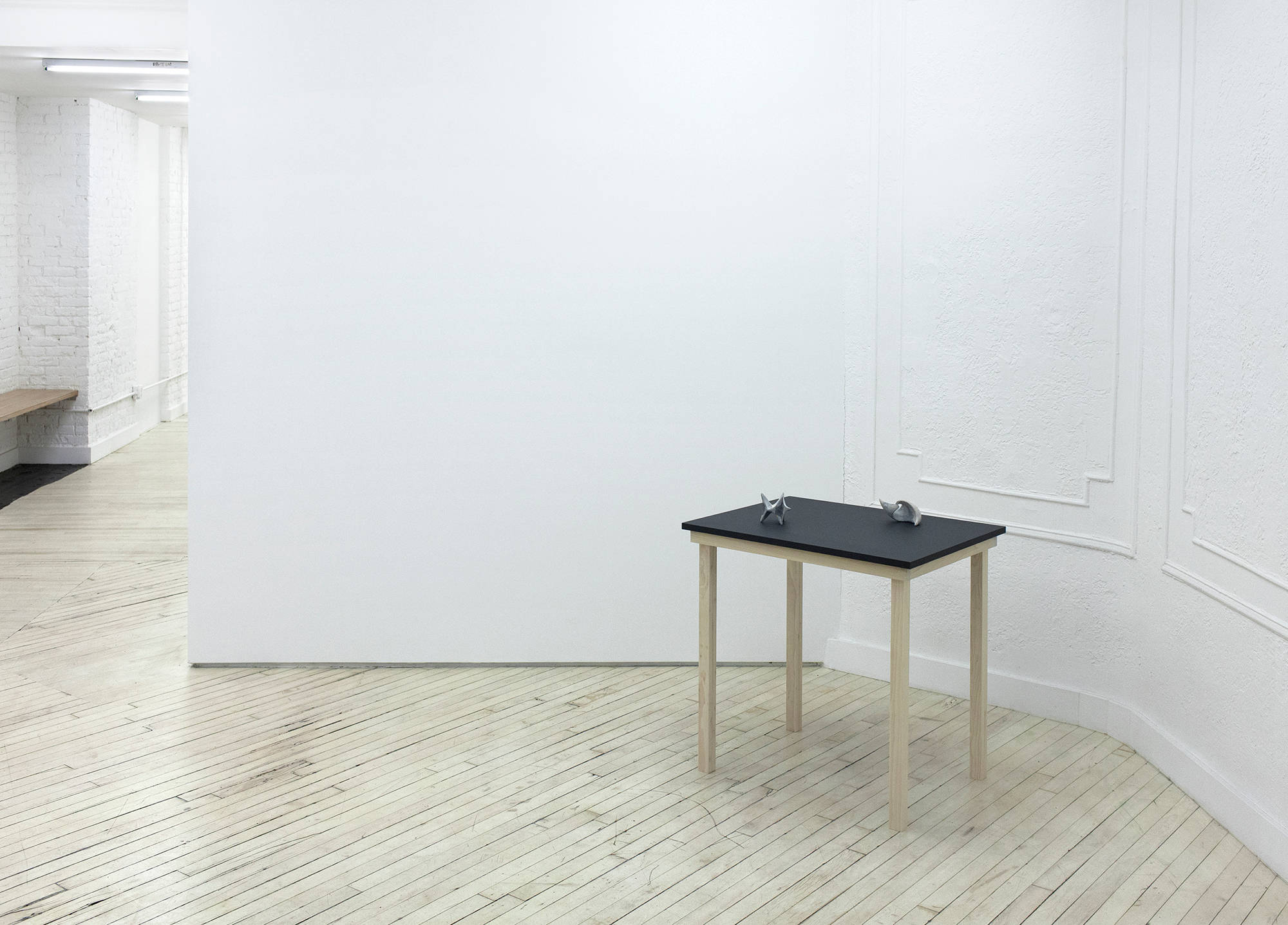 Installation view of a sculpture in a gallery by Matt Hoyt depicting a black table with wooden legs upon which two hand made sculptures sit.