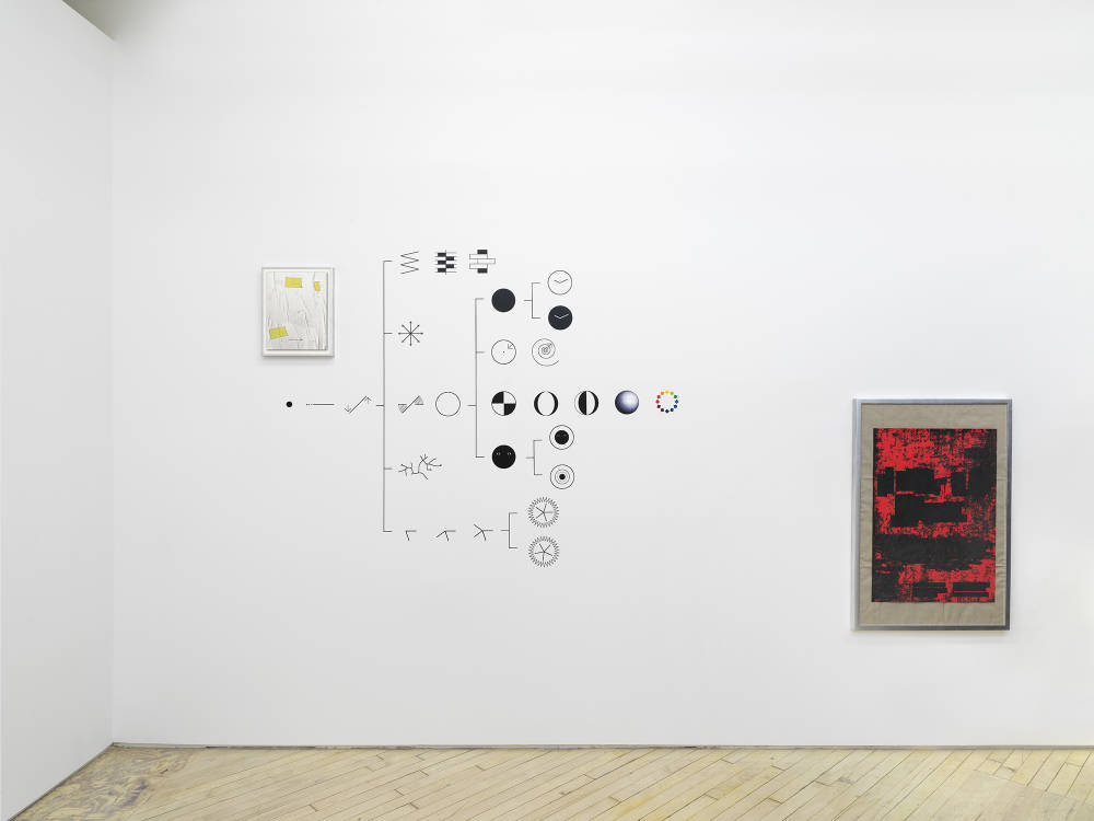 On a large wall, two graphic images are installed in frames at varying heights. In the center of the wall is a vinyl installation depicting the procession of a dot into varying symbols. 