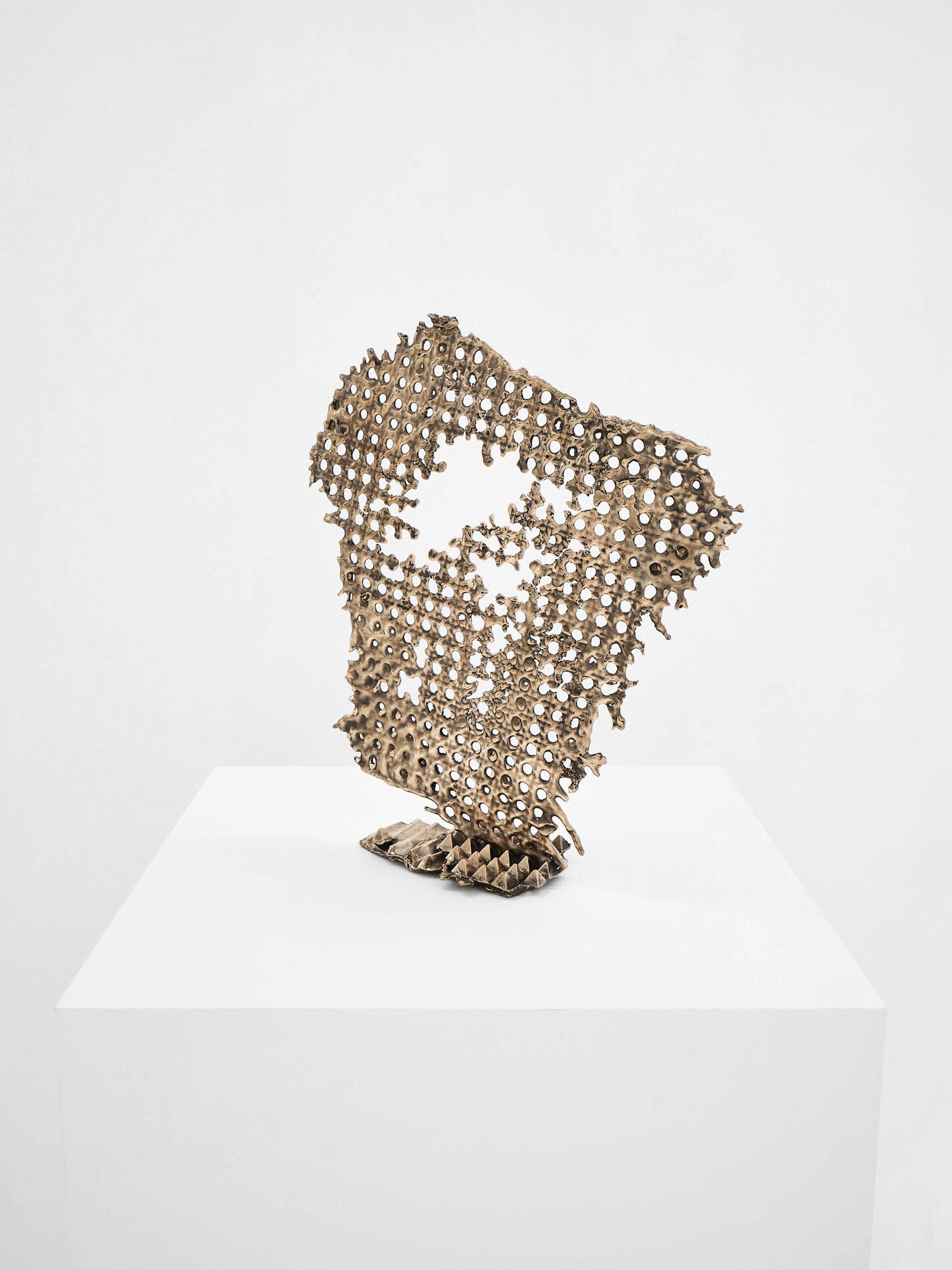 A bronze sculpture of cast chair cane sits on a white pedestal against a white wall.