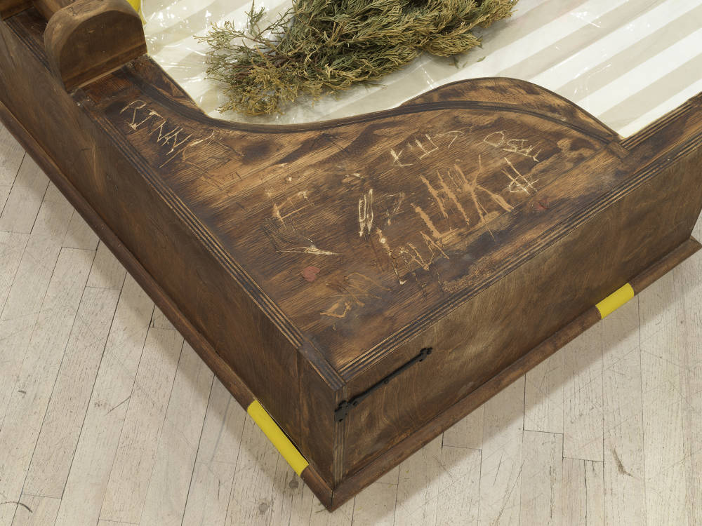 Image of an artwork by Libby Rothfeld of two conjoined boxes with yellow paint on the interior of the box. There is a dead Christmas tree in a circular cut out of the center of the box. The Christmas tree is embedded into what looks like a couch cushion with a plastic cover. There is graffiti scratched into the surface of the wooden box and bubblegum applied to it.