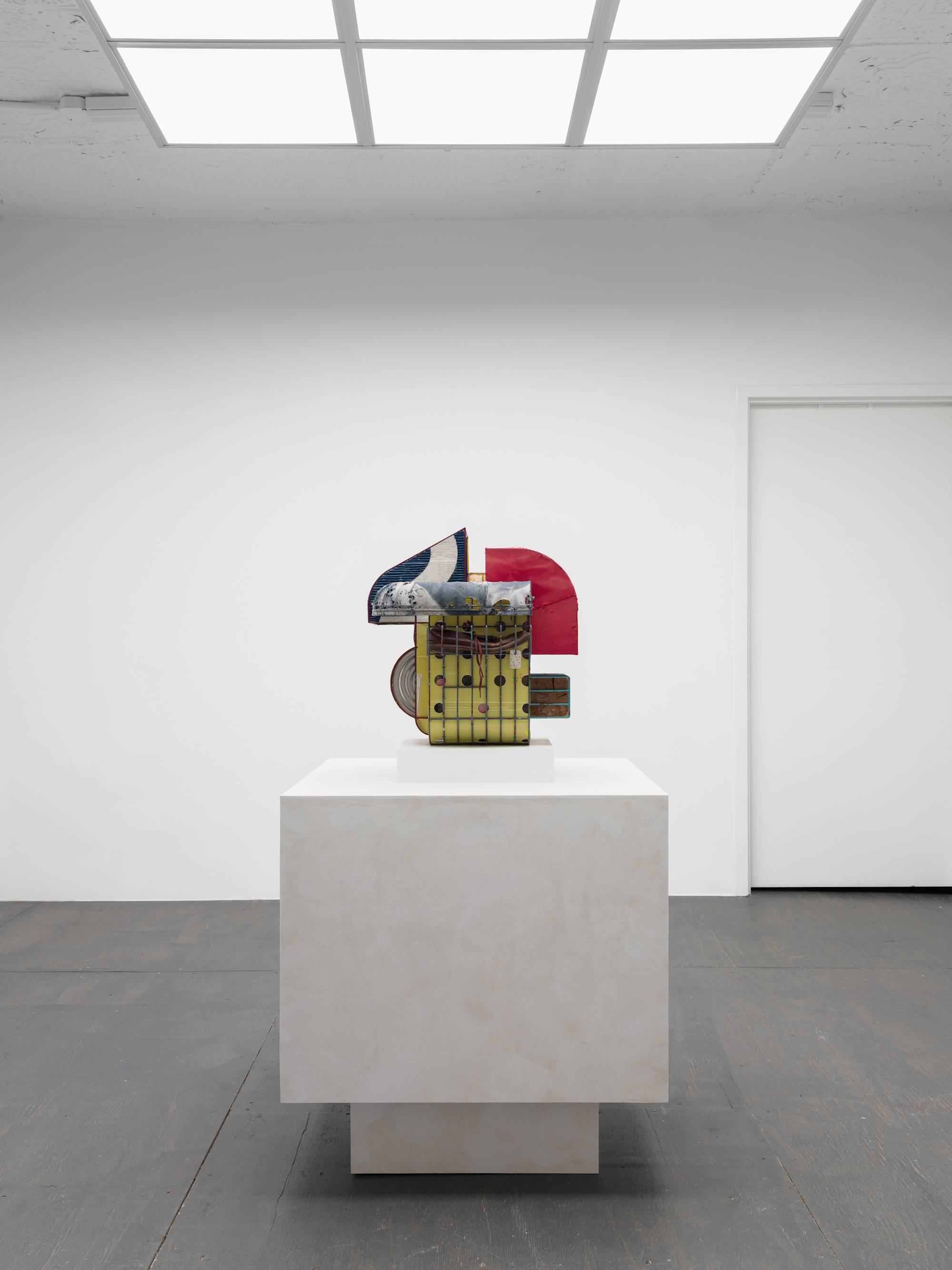 Straight on view of a colorful, cage-like sculpture, wrapped in tattered clothing, standing on a pedestal in the center of a white walled gallery with grey floor lit by an LED light fixture that resembles a skylight.