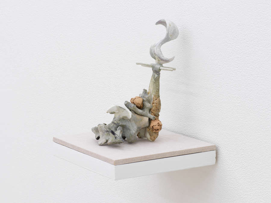 A small abstract sculpture resembling a kind of fossil, shell, bone formation, or combination of organic materials. The object is primarily gray and tan and resting on a shelf with a tan base attached to a white wall.