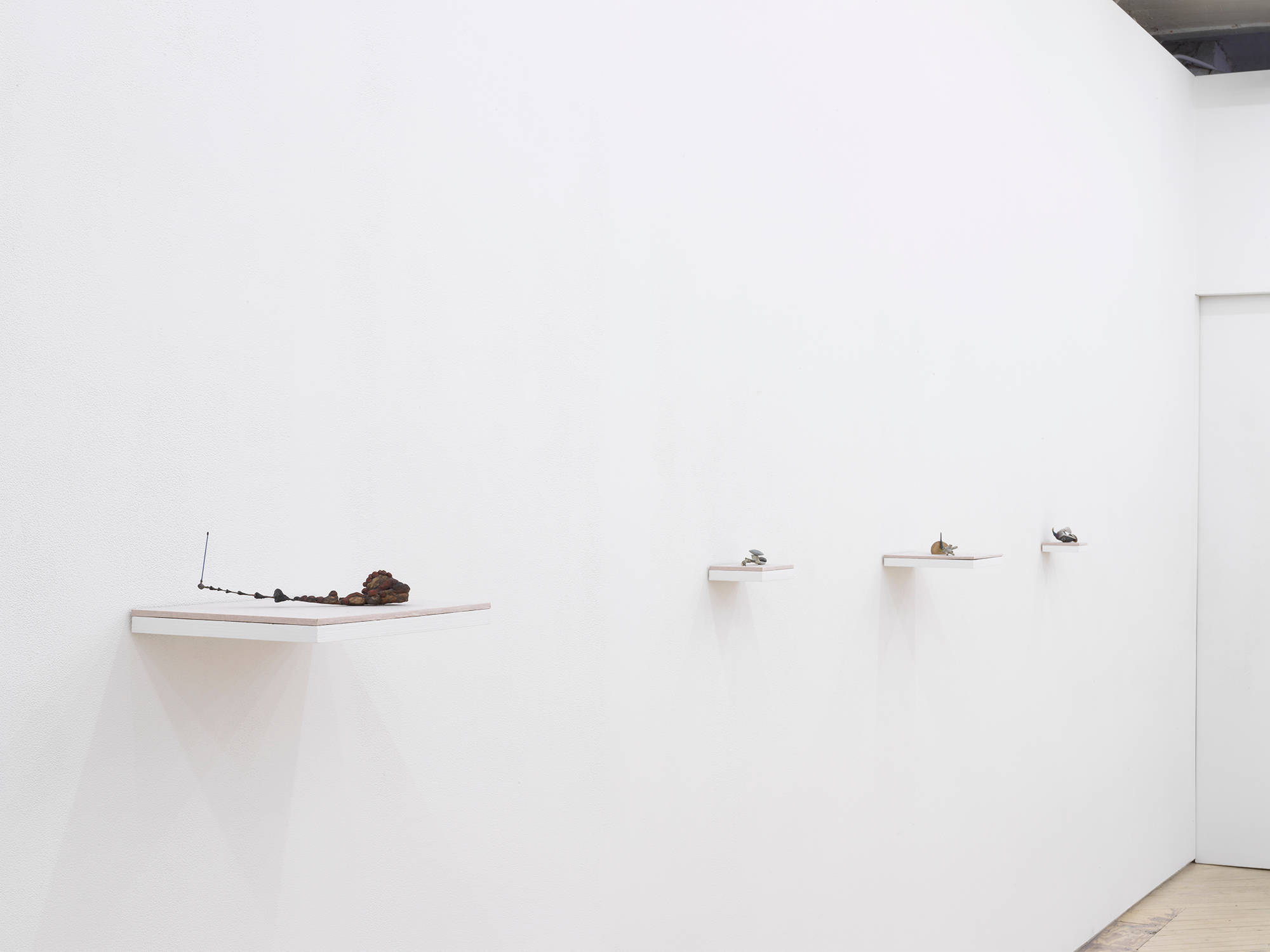 In a gallery space, a row of four miniature abstract sculptures placed on top of wall-mounted shelves evenly spaced apart.